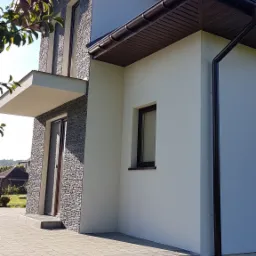 Nowoczesny dom jednorodzinny z elewacją łączącą biały tynk i kamienną okładzinę, brązowe rynny i stolarka okienna, widok z boku na taras wyłożony kostką.