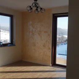 Wnętrze pokoju z dekoracyjnym tynkiem w odcieniach beżu, widok na zimowy krajobraz przez okno, nowoczesna lampa sufitowa.