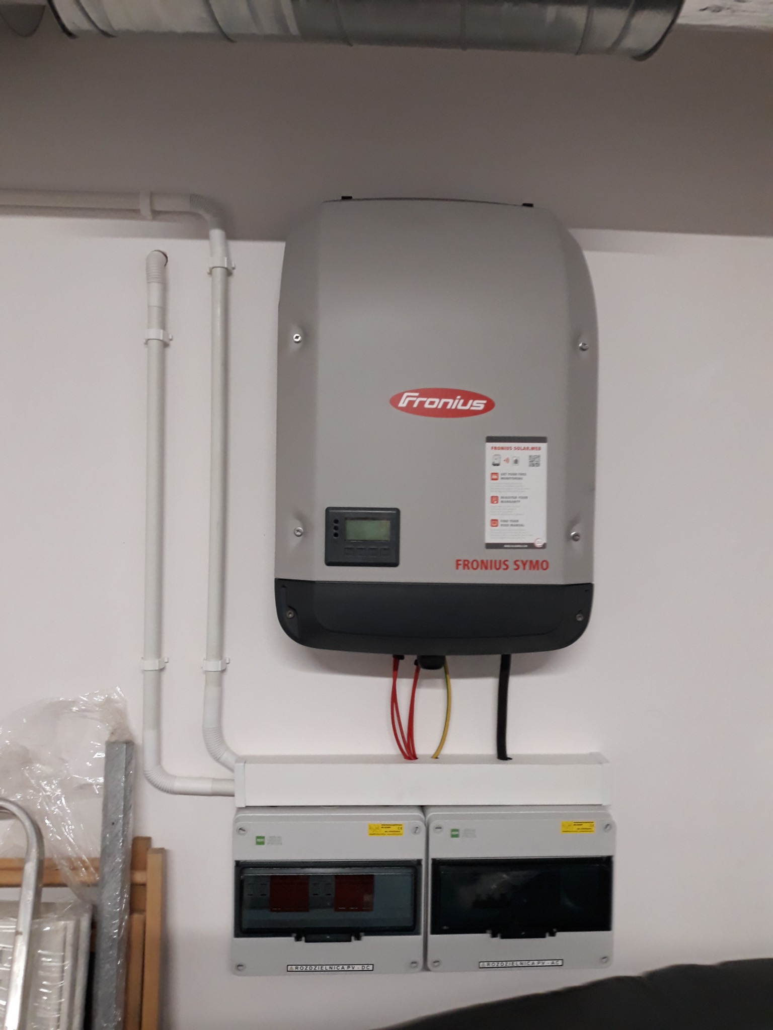 Szary inwerter Fronius Symo zamontowany na białej ścianie z widocznymi rurami instalacyjnymi i rozdzielnicami PV DC/AC poniżej.