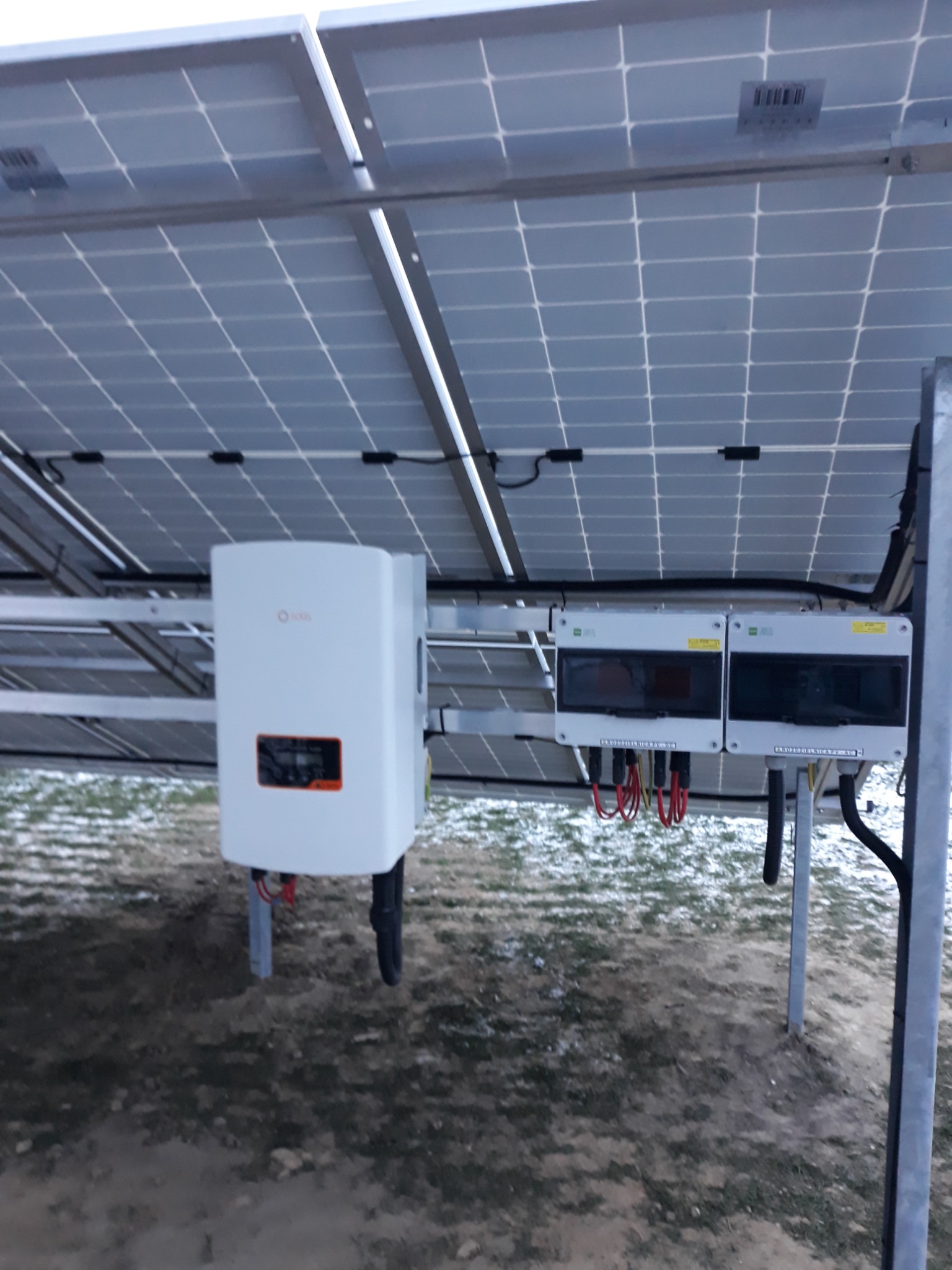 Widok od spodu na panele fotowoltaiczne, z widocznym inwerterem SOFAR i skrzynkami przyłączeniowymi, zamontowane na metalowej konstrukcji, na tle ziemi pokrytej resztkami śniegu.