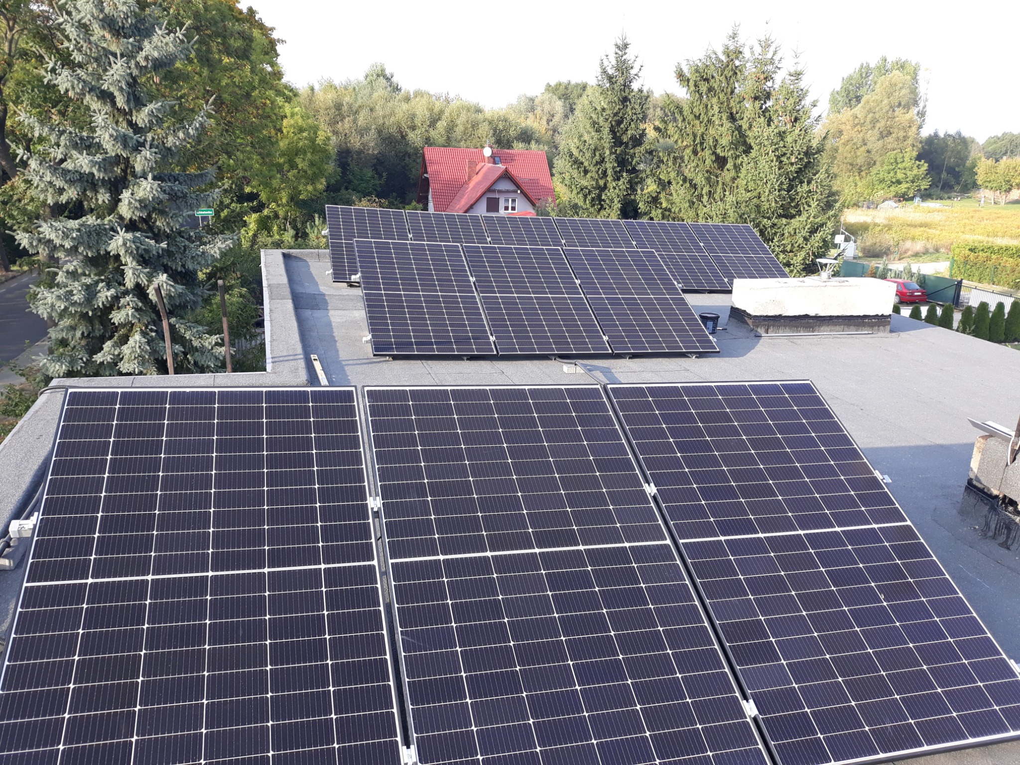 Instalacja paneli słonecznych na płaskim dachu domu jednorodzinnego, widok z góry, w tle drzewa i dom z czerwoną dachówką.