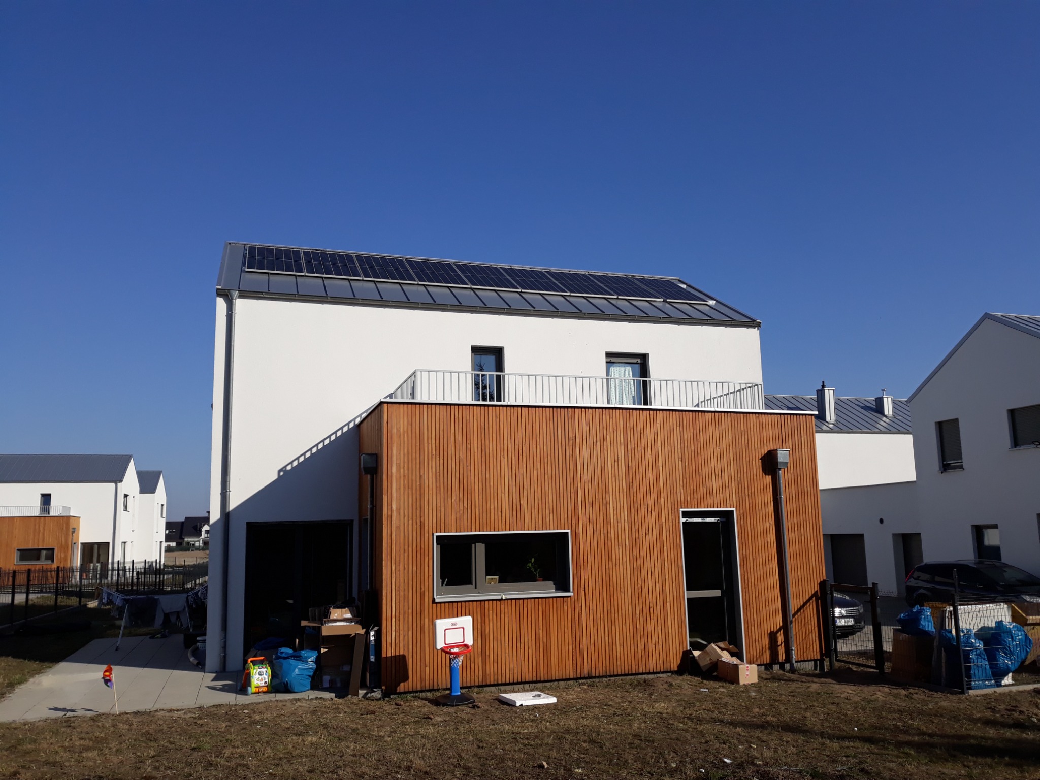 Nowoczesny dom jednorodzinny z panelami fotowoltaicznymi na dachu, drewnianą elewacją i balkonem z metalową barierką, widoczny mały kosz do koszykówki na trawniku.