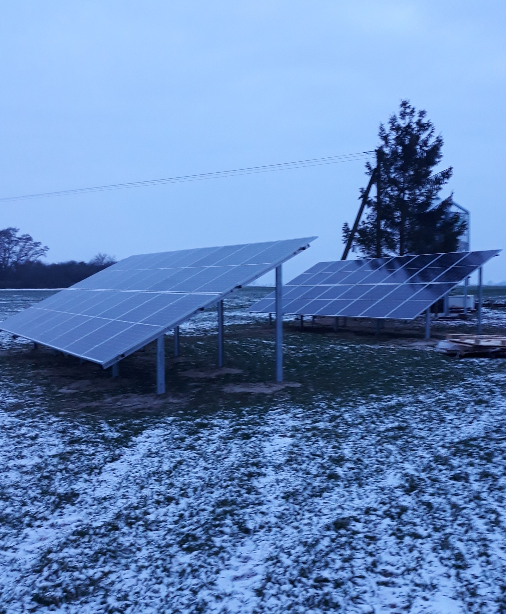 Dwa rzędy paneli słonecznych na metalowych konstrukcjach, częściowo pokryte śniegiem, na tle zimowego krajobrazu z drzewami i słupem energetycznym.