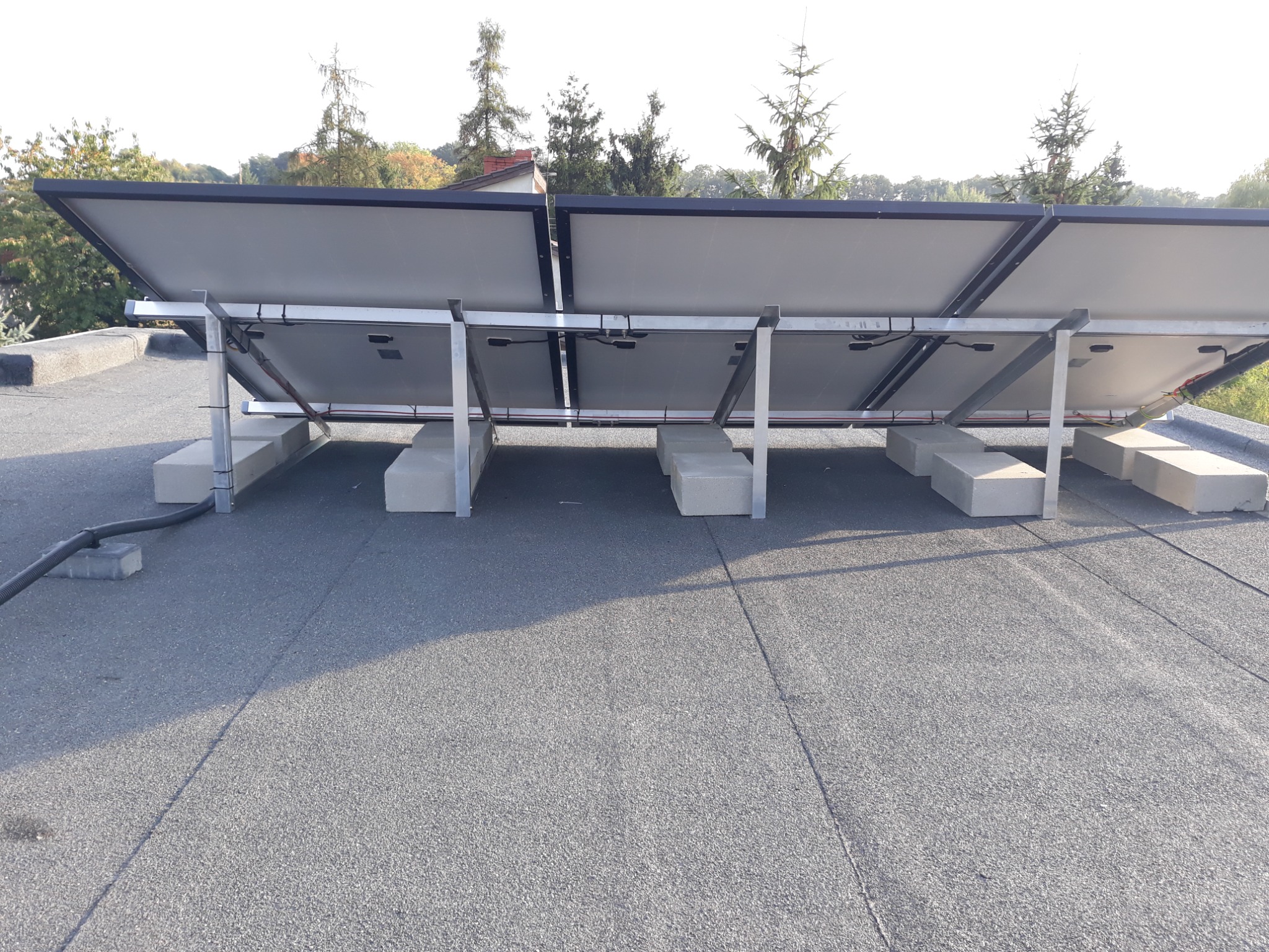 Montaż paneli słonecznych na płaskim dachu, widok od tyłu, z widocznymi betonowymi blokami podtrzymującymi konstrukcję i przewodami.