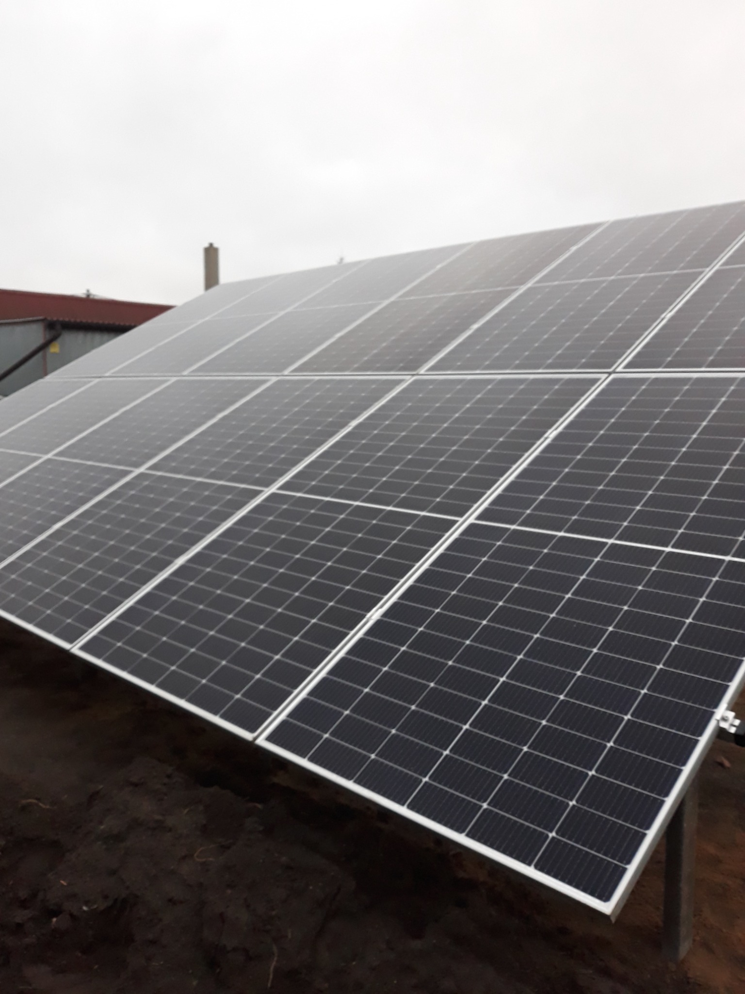 Panel fotowoltaiczny na gruncie, widoczne mocowania i fragment dachu budynku w tle, pochmurne niebo.