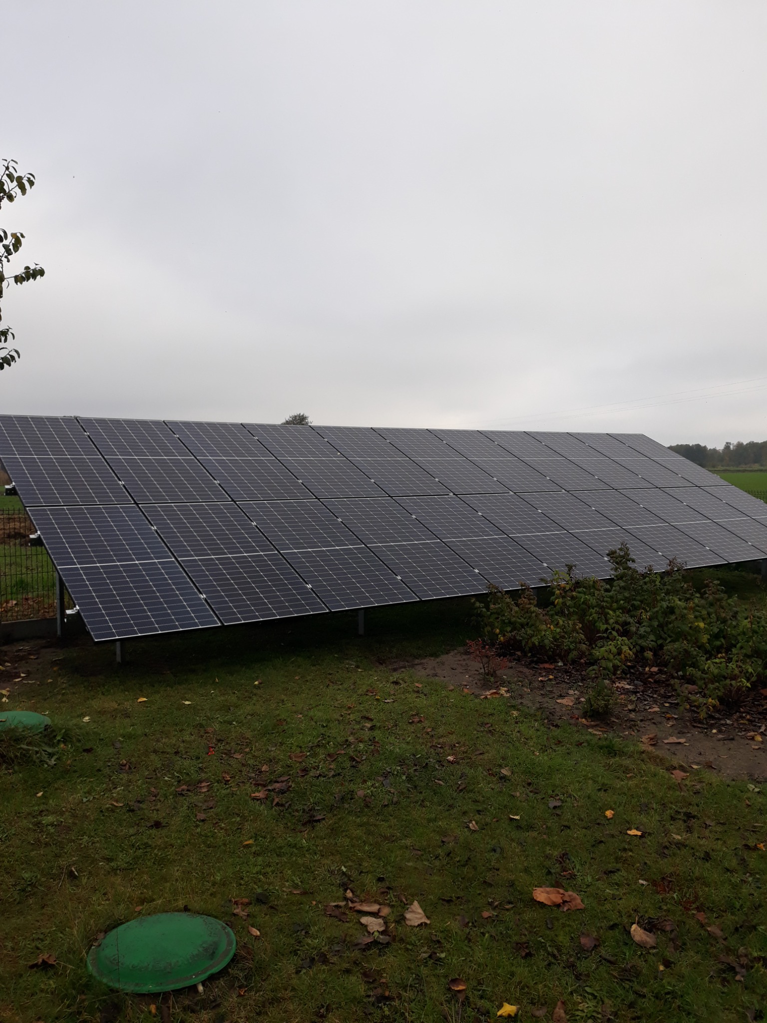 Rząd paneli słonecznych na metalowej konstrukcji, zamontowany na trawiastym terenie w pochmurny dzień, z widocznymi zielonymi pokrywami studzienek w trawie.