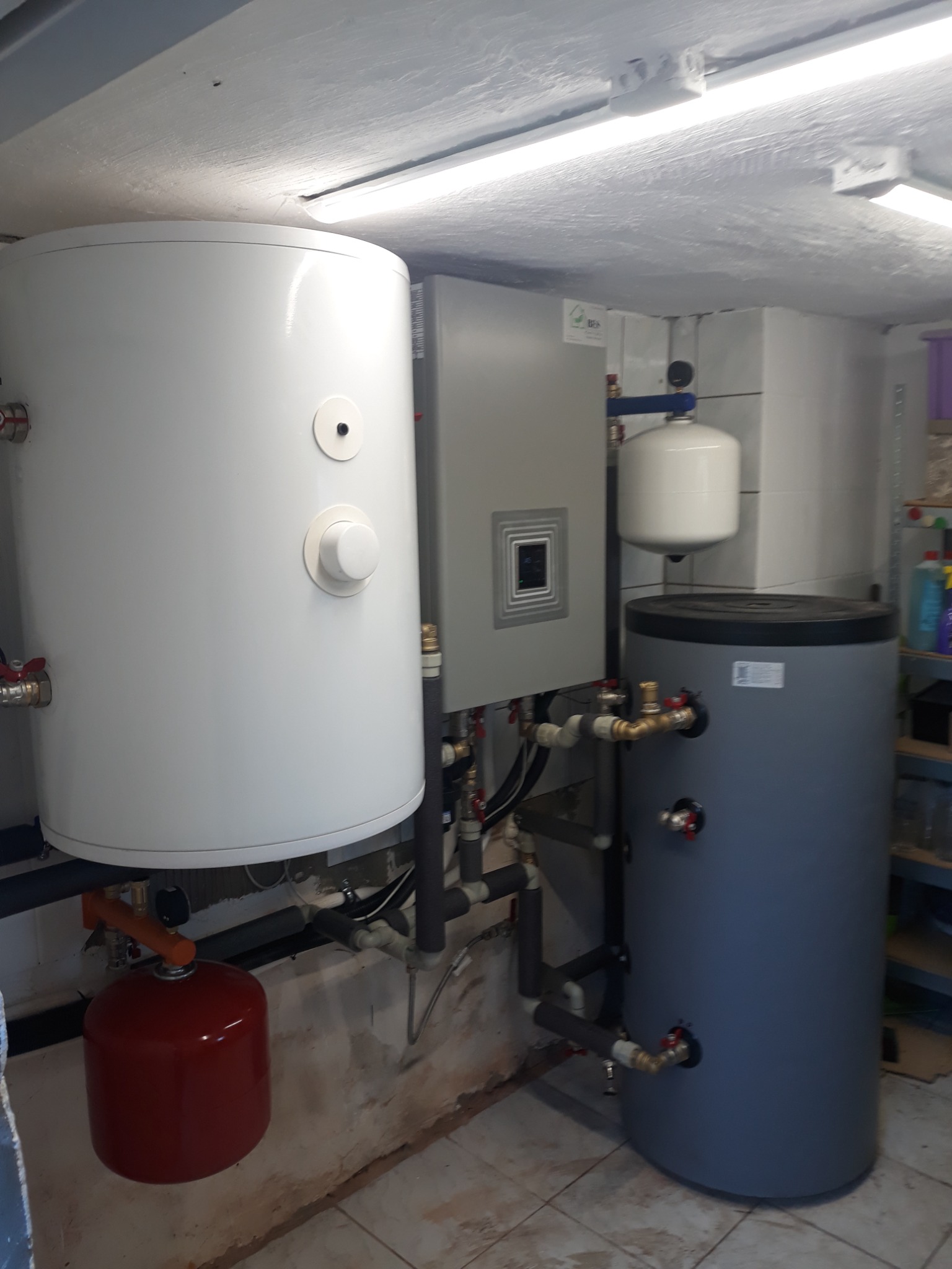 Kompletna instalacja grzewcza w piwnicy: biały zasobnik ciepłej wody, szary panel sterowania, szary zbiornik buforowy i czerwony zbiornik przeponowy, połączone rurami i zaworami.