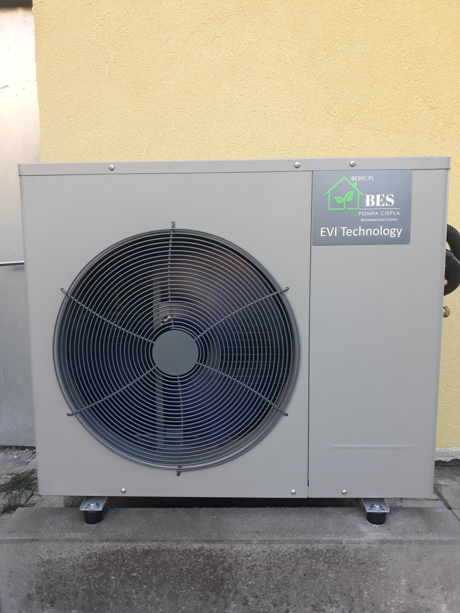 Szara pompa ciepła z wentylatorem na zewnątrz budynku, z logo firmy BES i informacją o technologii EVI, zamontowana na betonowym podłożu przy żółtej ścianie.