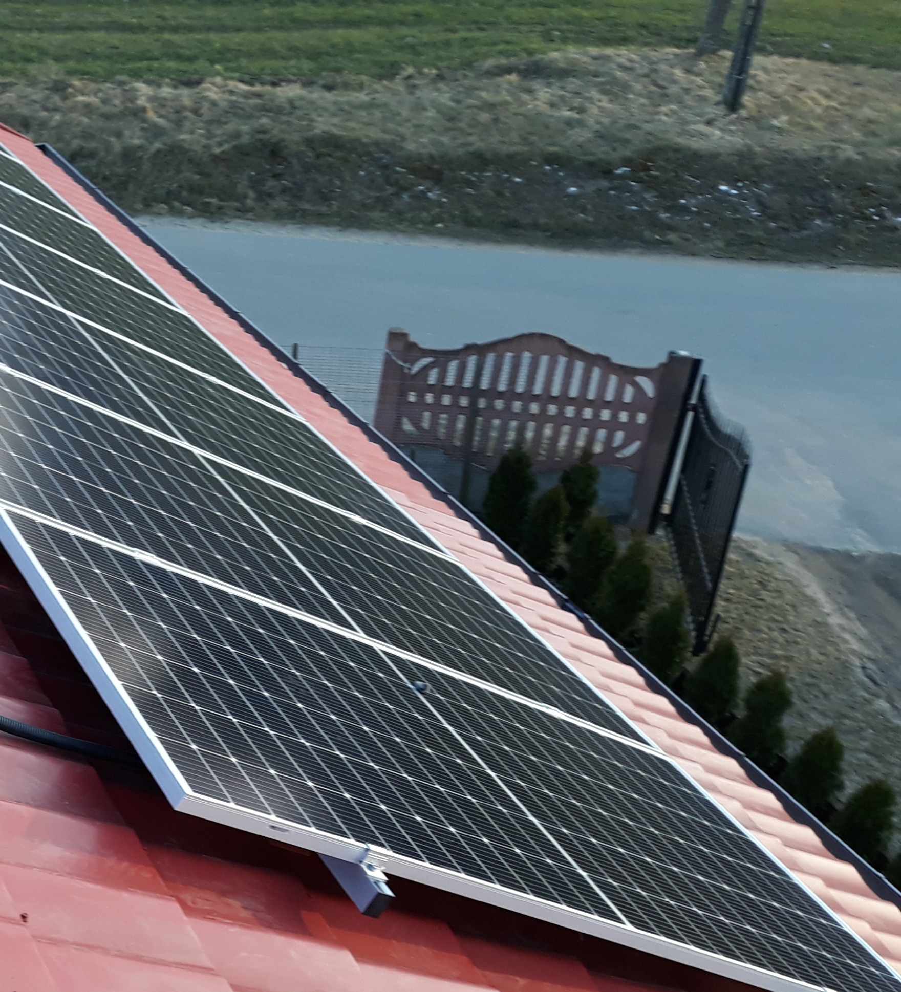 Panele słoneczne zamontowane na czerwonym dachu, widoczne elementy mocujące i przewody, w tle brama wjazdowa i zieleń.