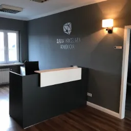 Recepcja w nowoczesnym biurze. Ciemnoszara ściana z białym napisem 'Śląska Kancelaria Adwokacka' i geometrycznym logo. Lampa ścienna, biała recepcja z czarnym frontem, widok na jasne pomieszczenie...