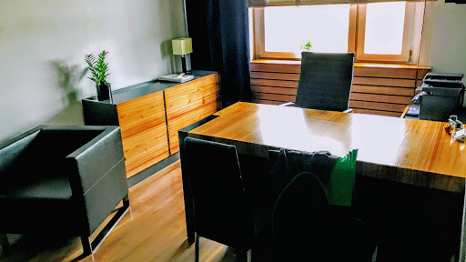 Wnętrze gabinetu z biurkiem z jasnego drewna, czarnymi krzesłami, komodą z rośliną doniczkową i oknem w tle.