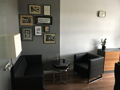 Wnętrze kancelarii: czarne skórzane sofy i fotel, stolik kawowy ze szklanym blatem, ściana ozdobiona ramkami z dyplomami i grafikami, zegar na ścianie, minimalistyczny design.