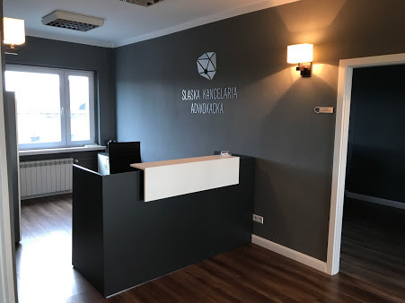 Recepcja w nowoczesnym biurze. Ciemnoszara ściana z białym napisem 'Śląska Kancelaria Adwokacka' i geometrycznym logo. Lampa ścienna, biała recepcja z czarnym frontem, widok na jasne pomieszczenie...