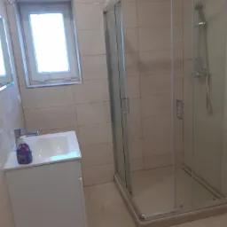 Jasna łazienka po renowacji z kabiną prysznicową z chromowanymi detalami, umywalką z szafką i oknem, wykończona beżowymi płytkami.