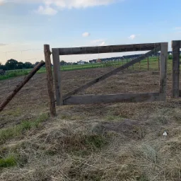 Drewniana brama z siatki leśnej na łące, w tle zabudowania mieszkalne i błękitne niebo.