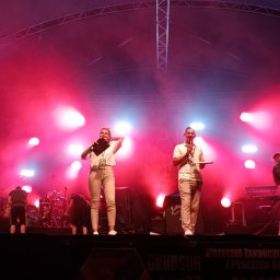 SELEDYNEK - Dwoje konferansjerów na scenie w czerwonym oświetleniu podczas wydarzenia plenerowego, w tle instrumenty muzyczne i ekipa techniczna.