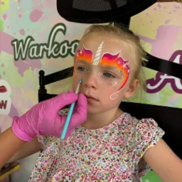 Dziewczynka z pomalowaną twarzą w motywie jednorożca, błyszczący brokat i gradient kolorów, malowana przez osobę w różowych rękawiczkach, na tle kolorowej ściany.