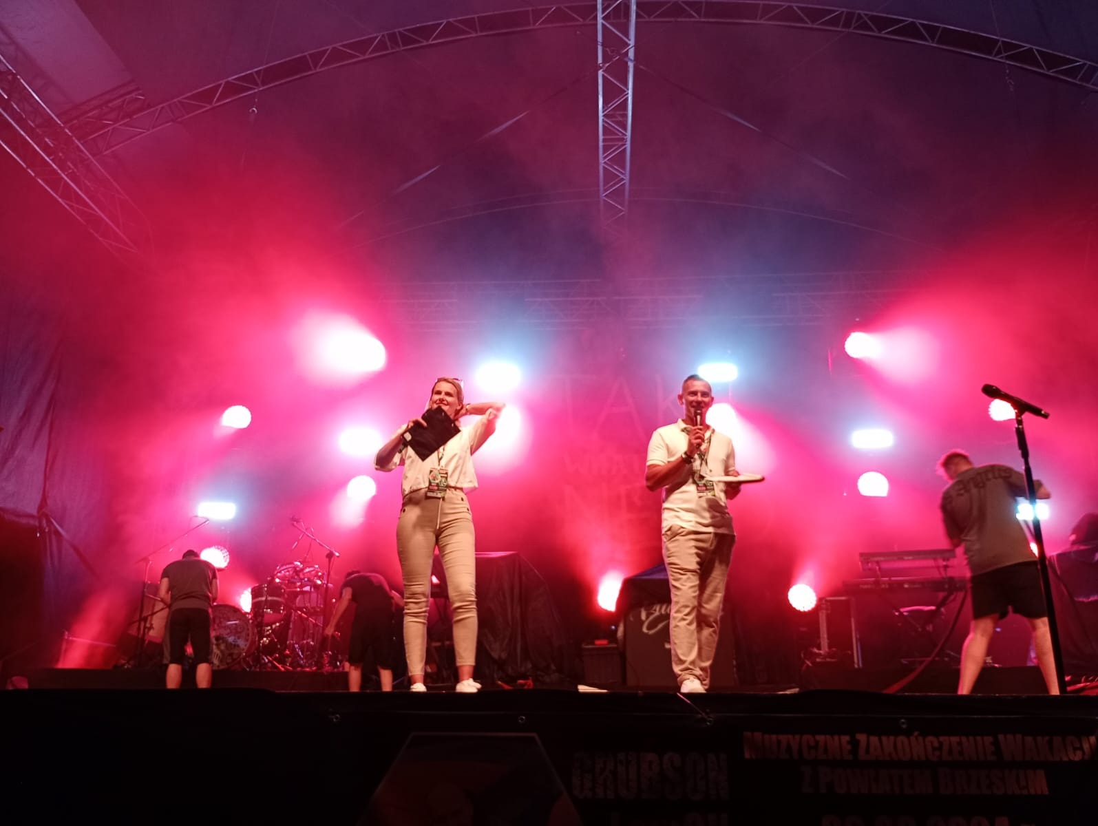 Dwoje konferansjerów na scenie w czerwonym oświetleniu podczas wydarzenia plenerowego, w tle instrumenty muzyczne i ekipa techniczna.