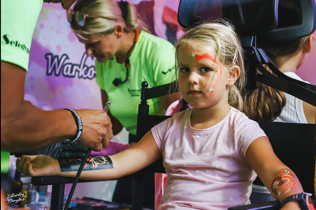 Dziewczynka z pomalowaną twarzą i wzorem na ramieniu, wykonywanym aerografem przez artystę, w tle inne osoby i logo firmy.