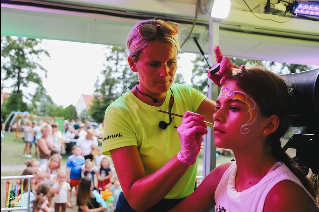 Kobieta w lateksowych rękawiczkach maluje twarz dziewczynki wzorem korony i chmurki na tle plenerowej imprezy.