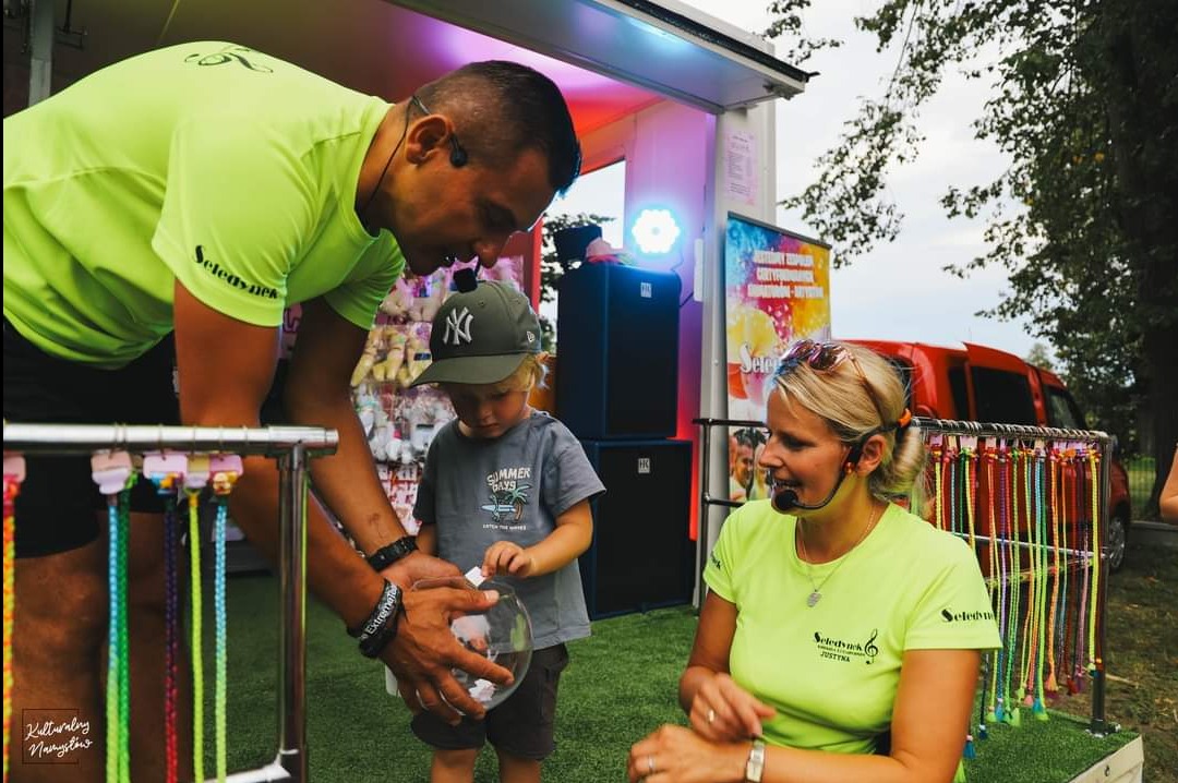 Animatorzy w neonowych koszulkach z mikrofonami podczas pracy: mężczyzna pomaga dziecku w losowaniu nagrody z przezroczystej kuli, a kobieta uśmiecha się w tle, widoczne kolorowe sznurki...