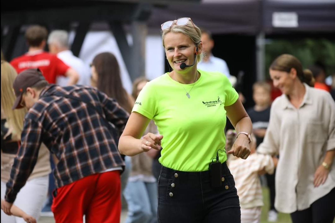 Uśmiechnięta animatorka w neonowej koszulce z logo, mikrofonem nagłownym i okularami na głowie prowadzi zabawę z dziećmi na imprezie plenerowej.