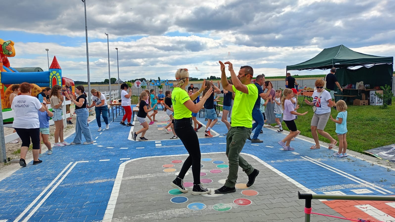 Animatorzy w neonowych koszulkach prowadzą gry i zabawy dla dzieci na zewnątrz, na placu zabaw z namalowanymi elementami gier planszowych i dmuchanym zamkiem w tle.
