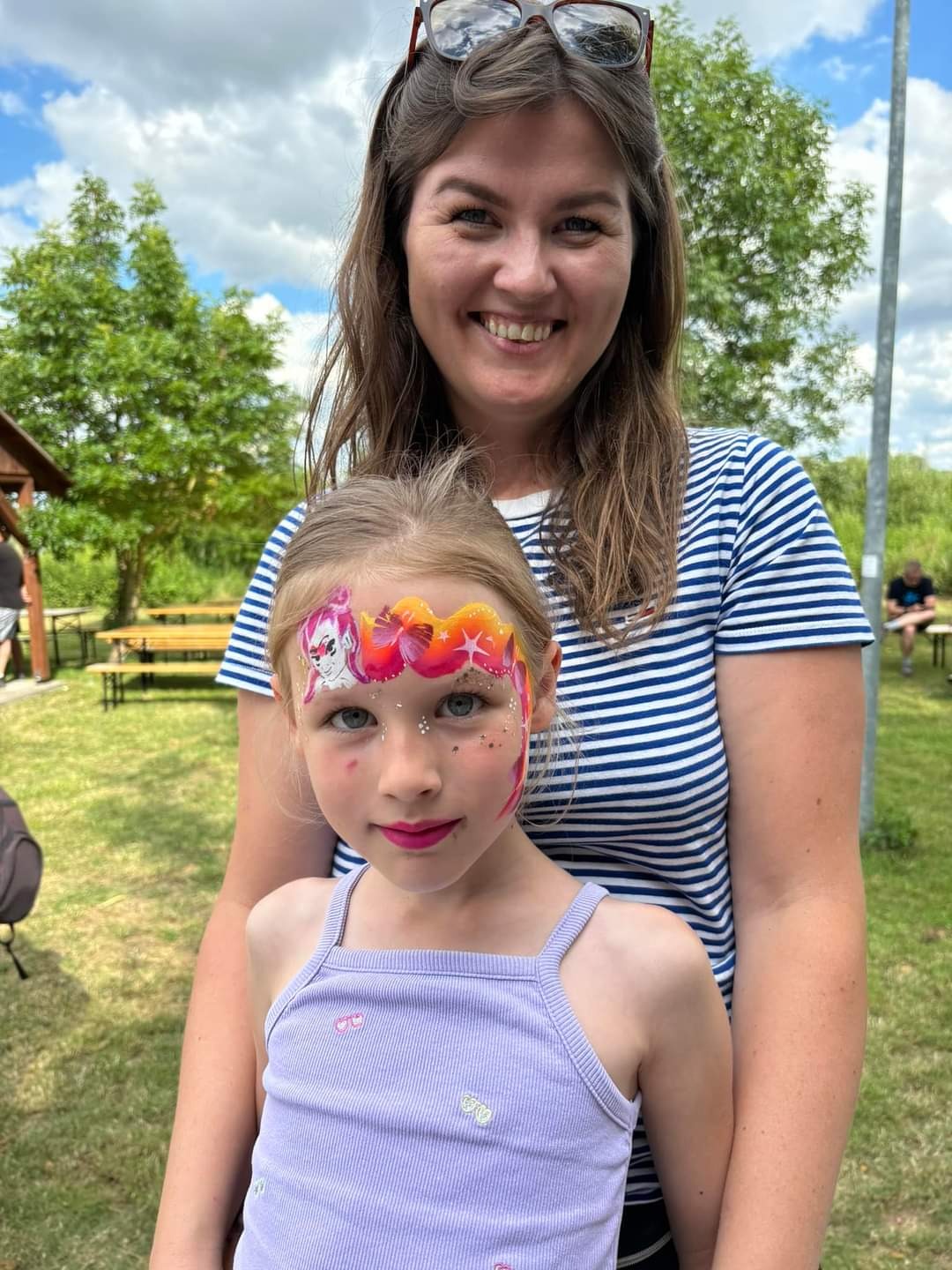 Dziewczynka z pomalowaną twarzą w stylu Paw Patrol, trzymana przez uśmiechniętą kobietę na tle zielonego trawnika i drzew.