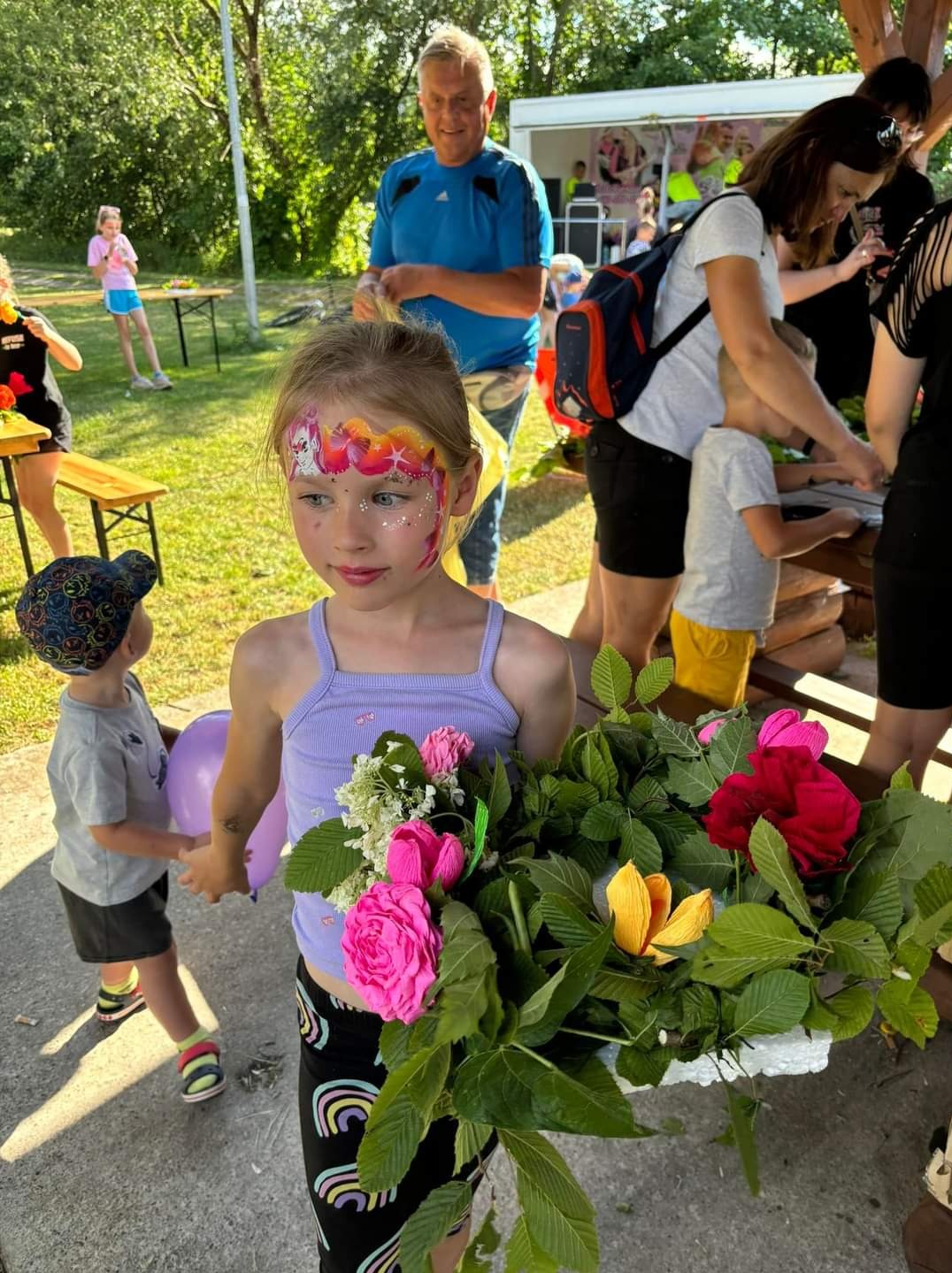 Dziewczynka z pomalowaną twarzą w motywy jednorożca trzyma bukiet z różowych i czerwonych róż oraz żółtego kwiatu, w tle widoczni inni uczestnicy imprezy plenerowej.