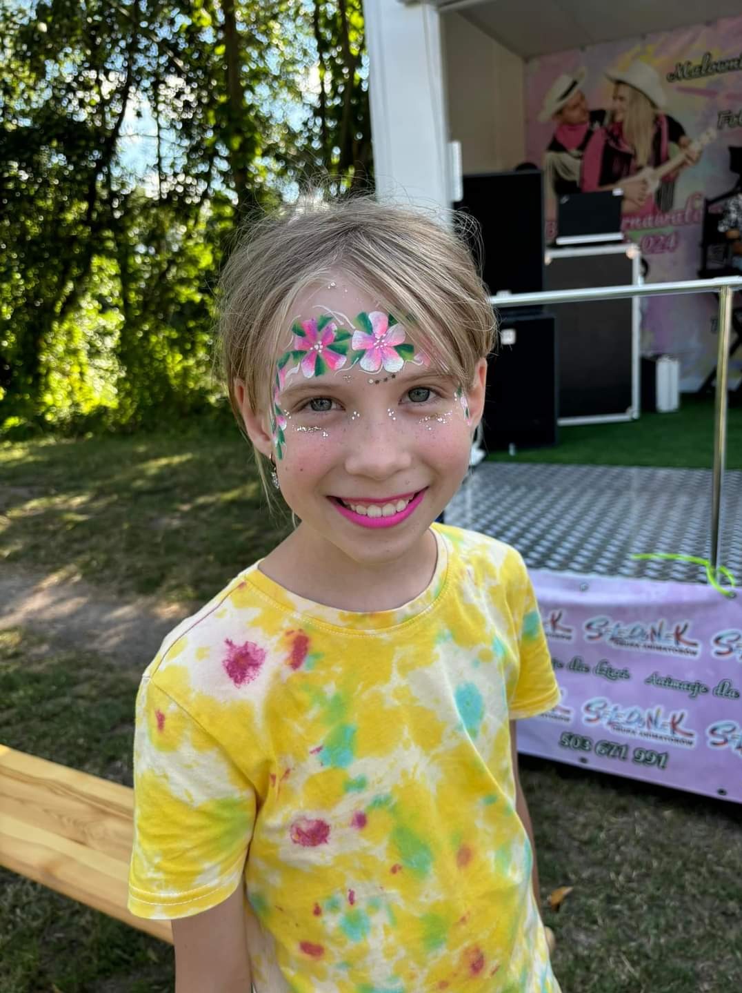 Uśmiechnięta dziewczynka z kwiatowym makijażem na twarzy, stojąca na tle zieleni i sceny z zespołem muzycznym na plakacie.