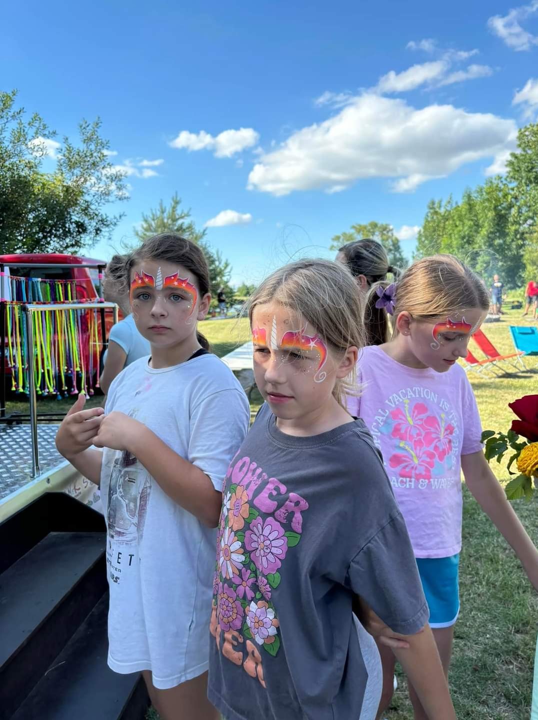 Trzy dziewczynki z pomalowanymi twarzami w motywie jednorożca, stojące obok wózka z kolorowymi wstążkami na tle zieleni i błękitnego nieba z chmurami.