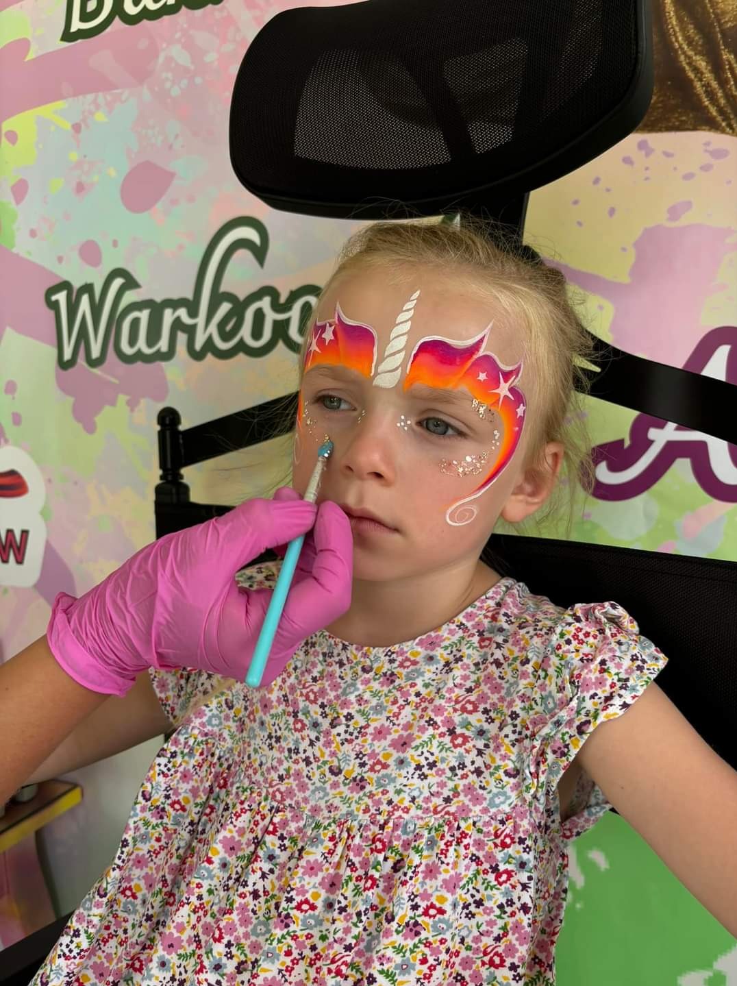 Dziewczynka z pomalowaną twarzą w motywie jednorożca, błyszczący brokat i gradient kolorów, malowana przez osobę w różowych rękawiczkach, na tle kolorowej ściany.