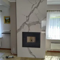 Częściowo wykończony kominek z płyt imitujących marmur, czarny wkład kominkowy, widoczne narzędzia glazurnicze na podłodze z płytek ułożonych w jodełkę.