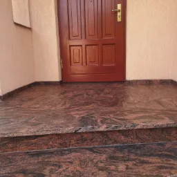 Wykończone wejście do domu w Jurowcach z brązowo-czerwonymi stopniami i podestem z materiału imitującego kamień, dopasowanymi do listew przypodłogowych, przed drewnianymi drzwiami.