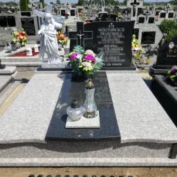 Granitowy nagrobek z czarną tablicą, ozdobiony kwiatami i lampionami, na tle cmentarza z licznymi grobami