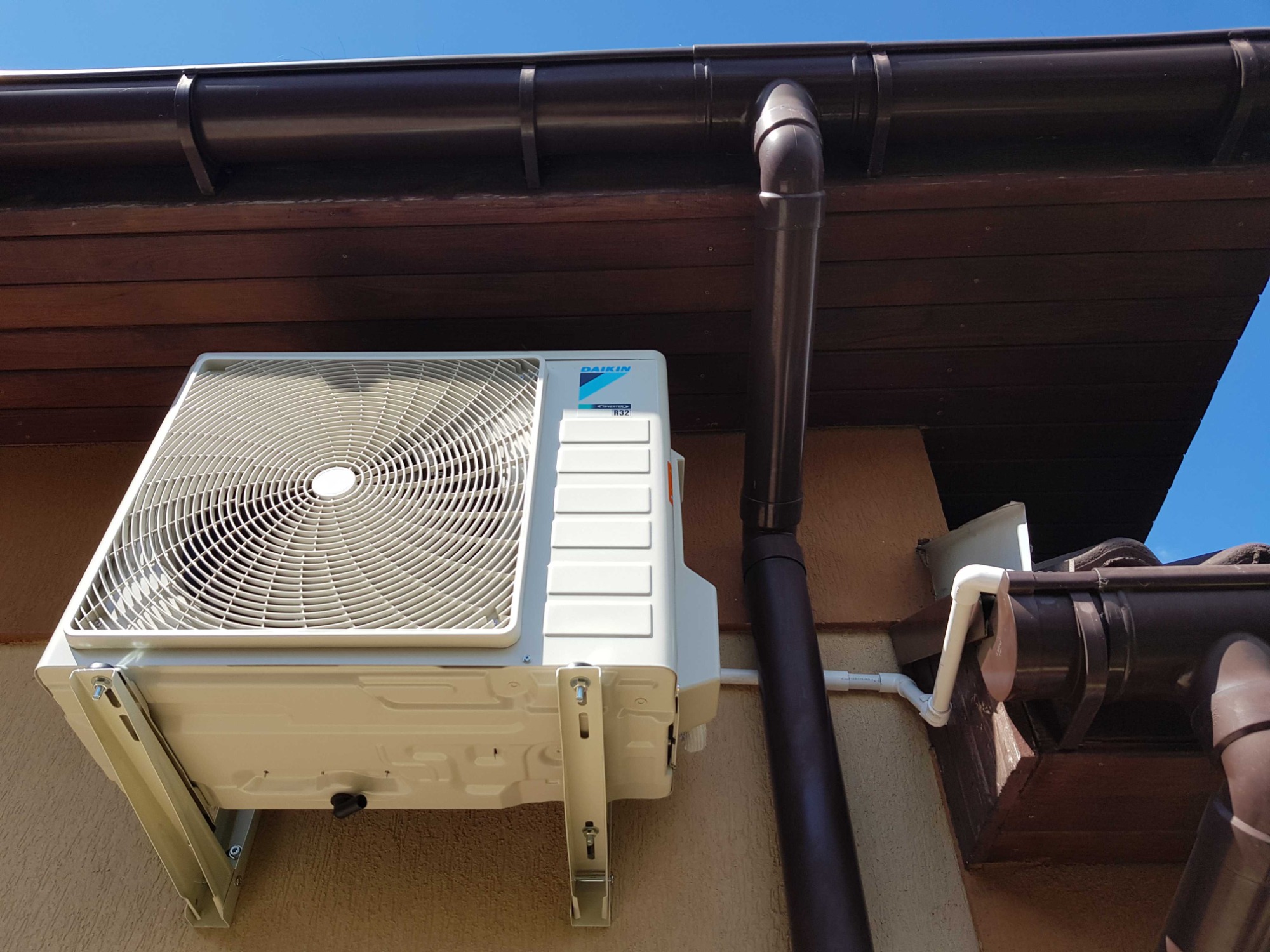 Zewnętrzna jednostka klimatyzacji Daikin zamontowana na ścianie budynku, widoczny brązowy system rynnowy i fragment niebieskiego nieba.