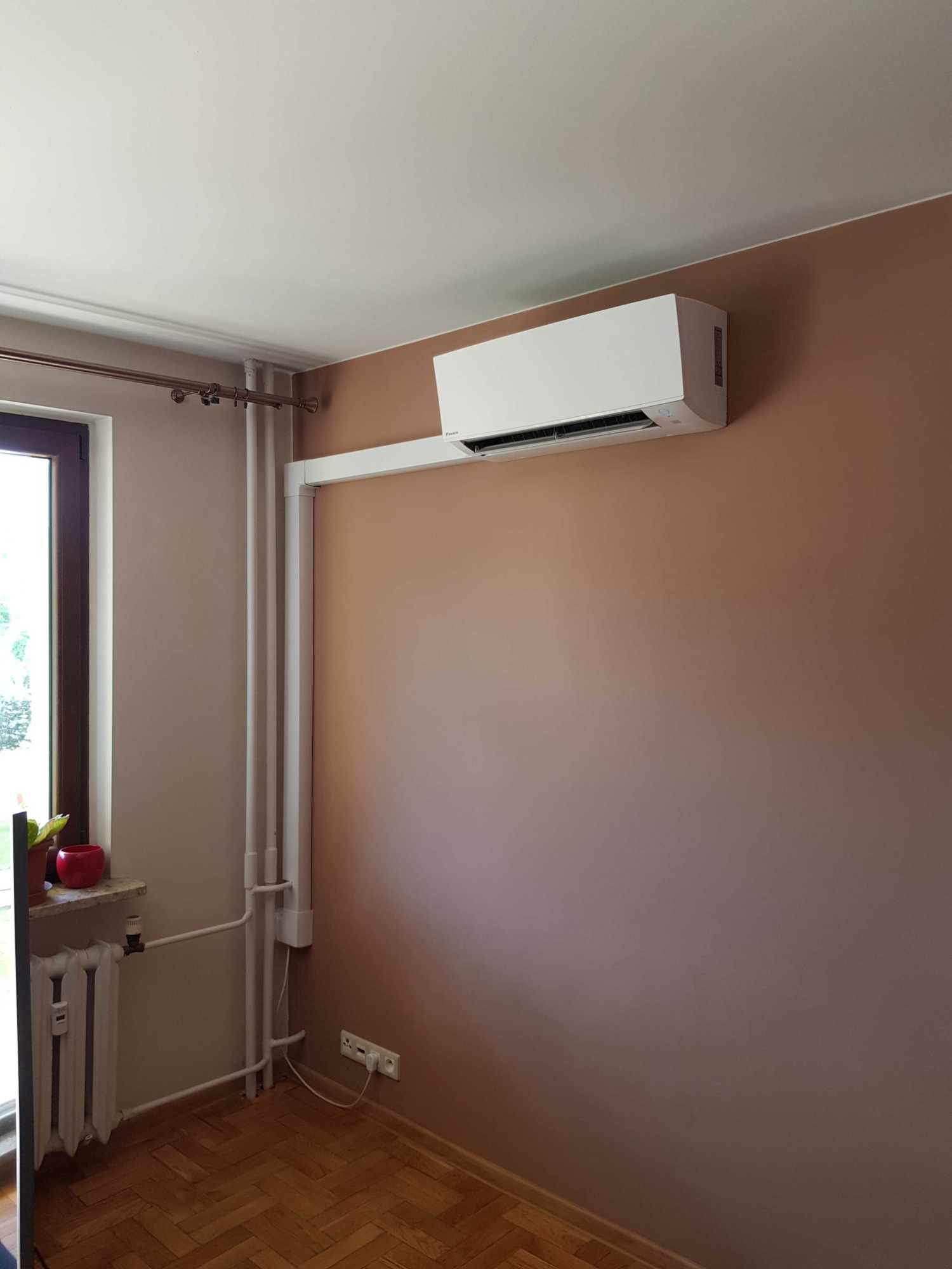 Biała jednostka klimatyzacji zamontowana na brązowej ścianie w pokoju z parkietem, widoczne rury i grzejnik przy oknie.