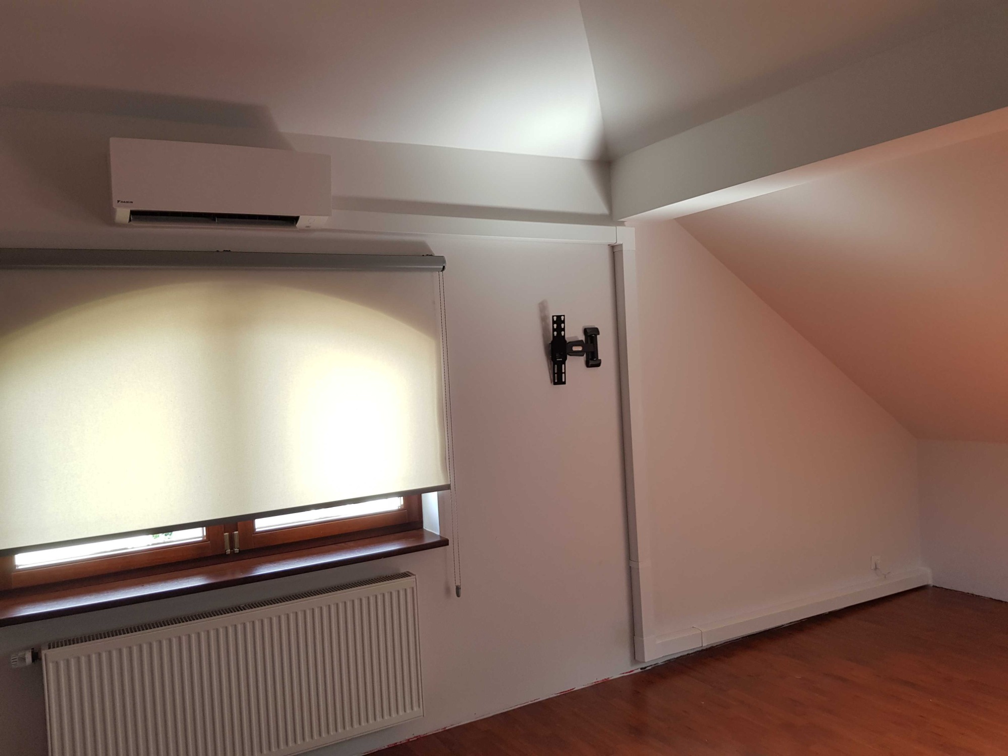 Biała jednostka klimatyzacji Daikin zamontowana pod sufitem w pokoju na poddaszu, widoczny grzejnik pod oknem z roletą, oraz uchwyt do telewizora na ścianie.