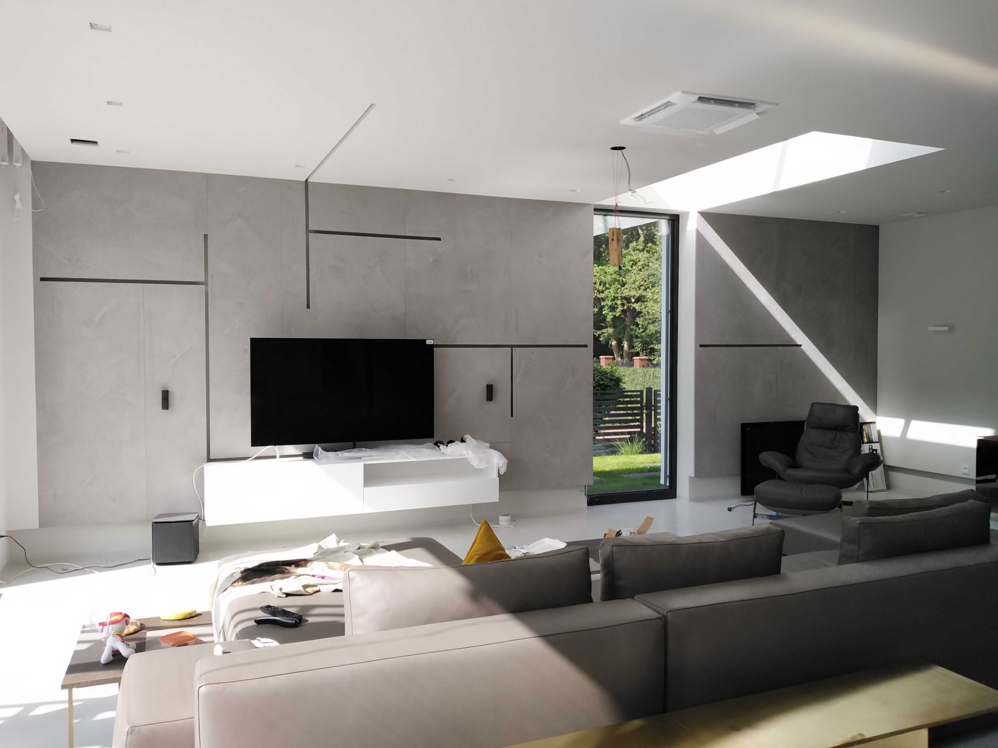 Wnętrze salonu w trakcie prac wykończeniowych, widoczny modułowy narożnik, fotel z podnóżkiem, wisząca szafka RTV, sufit podwieszany z klimatyzatorem kasetonowym i oknem dachowym.