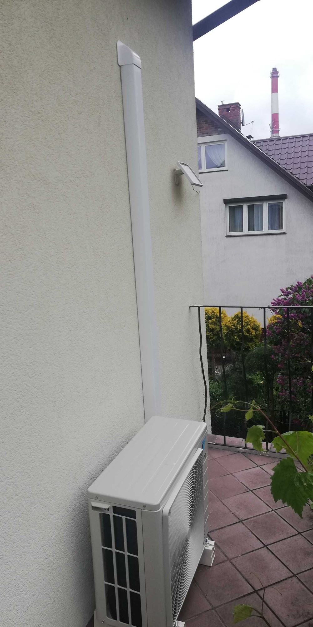 Zewnętrzna jednostka klimatyzacji zamontowana na balkonie domu jednorodzinnego, z widoczną białą rurą odprowadzającą skropliny wzdłuż ściany, w tle ogród i komin przemysłowy.