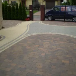 Kompozycja z wielobarwnej kostki brukowej na podjeździe w Jaworznie, zarysowana linia oddzielająca kolory, brama w tle z zaparkowanym vanem.