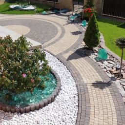 Styl-Bruk - Ogród z krętą ścieżką z kostki brukowej w odcieniach beżu i szarości, ozdobiony białymi kamykami, szkłem dekoracyjnym i roślinnością, w słoneczny dzień.