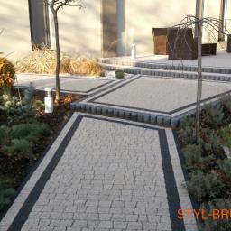 Styl-Bruk - Ogród z jasnoszarą kostką z ciemnymi obramowaniami, obrzeża z szarej kostki, rośliny ozdobne wzdłuż ścieżki, nowoczesny dom w tle, oświetlenie ogrodowe.