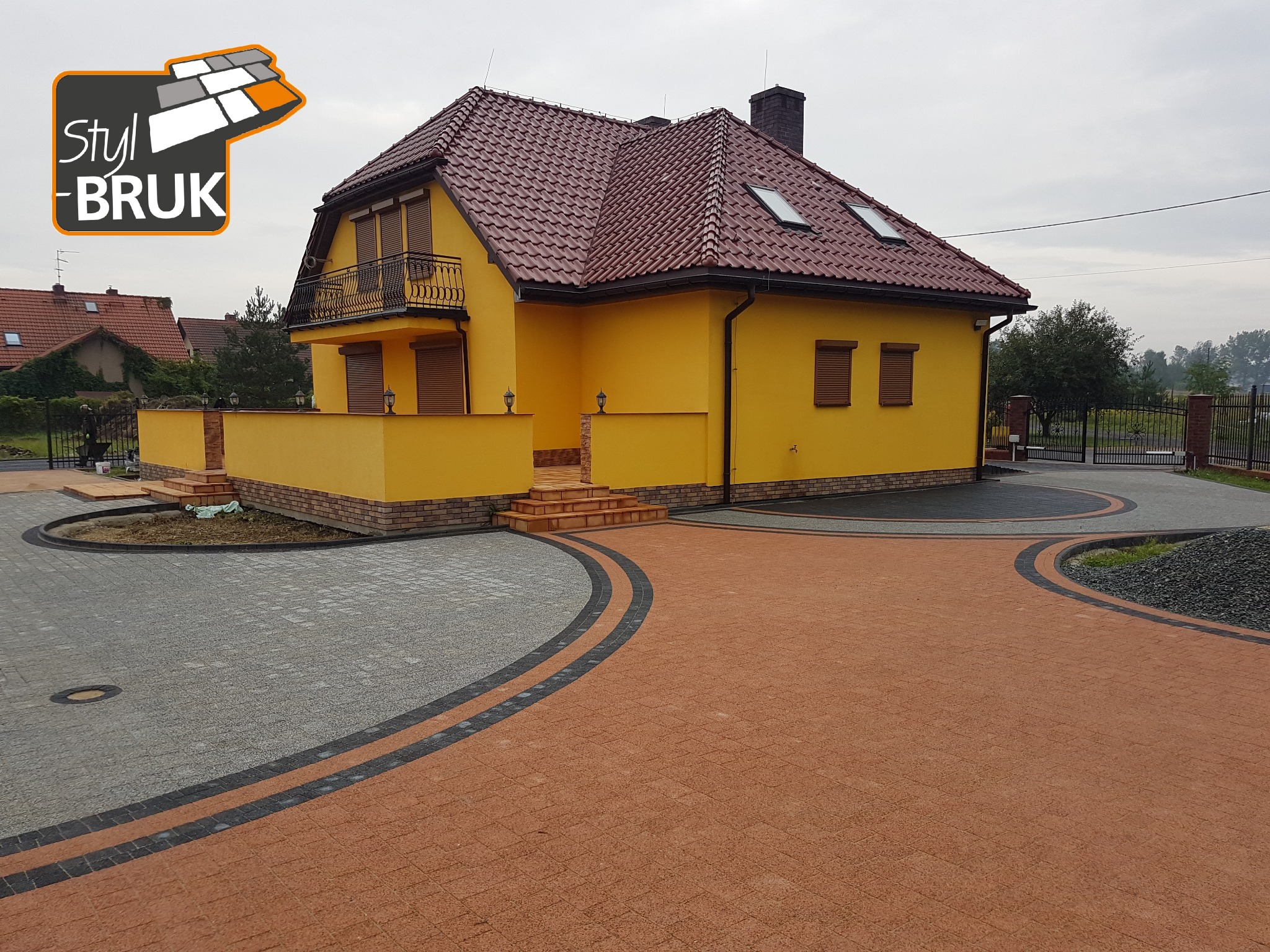 Żółty dom jednorodzinny z brązowym dachem i ozdobną, wielobarwną nawierzchnią z kostki brukowej, z logo firmy brukarskiej w lewym górnym rogu.