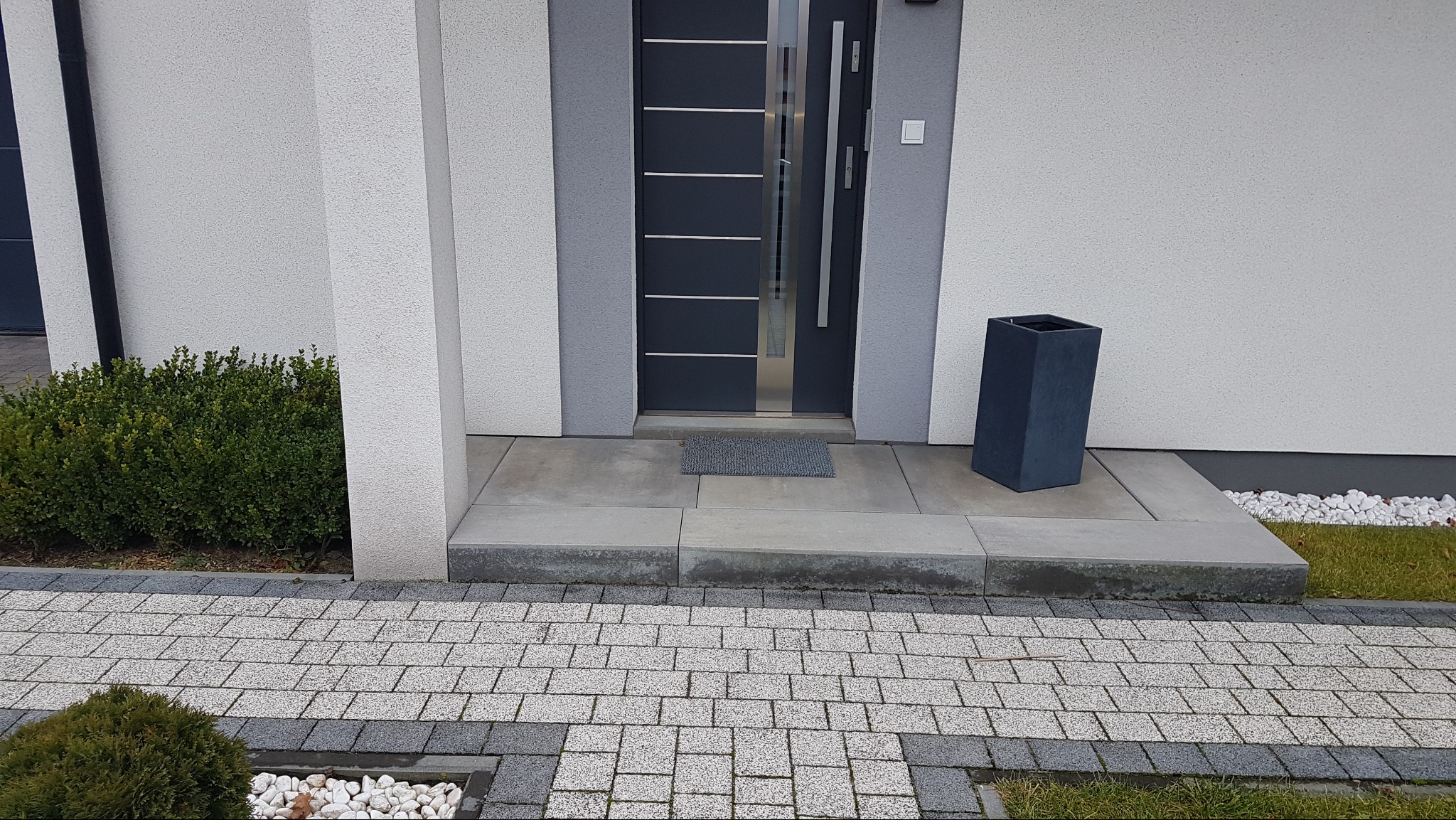Nowoczesne wejście do domu z szarej kostki brukowej, z obrzeżami z ciemniejszej kostki, prowadzące do betonowego podestu przed drzwiami i minimalistycznej donicy.
