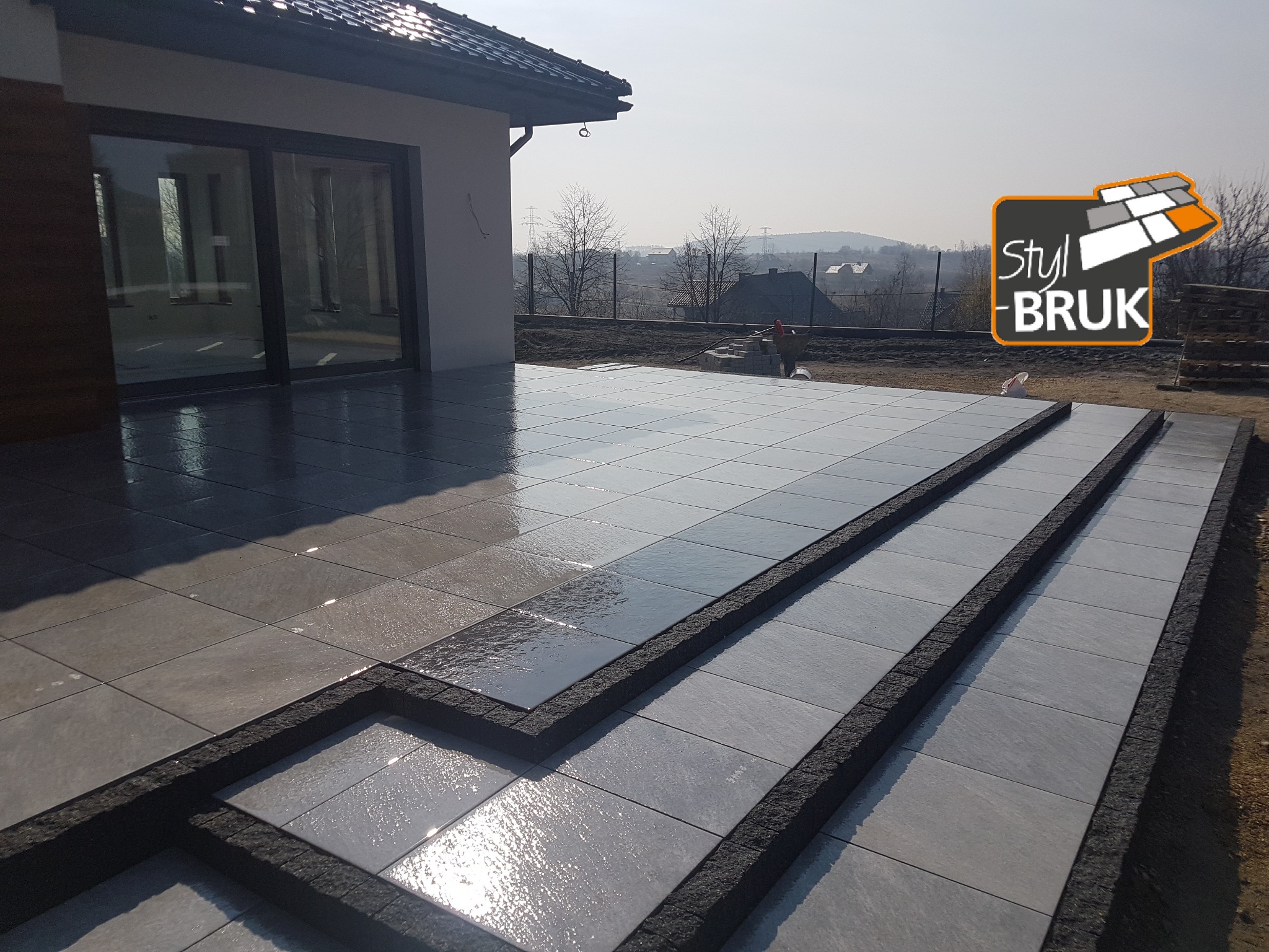 Nowoczesny taras z szarych płyt betonowych ułożonych schodkowo, z widocznym logo firmy brukarskiej w prawym górnym rogu i domem w tle.