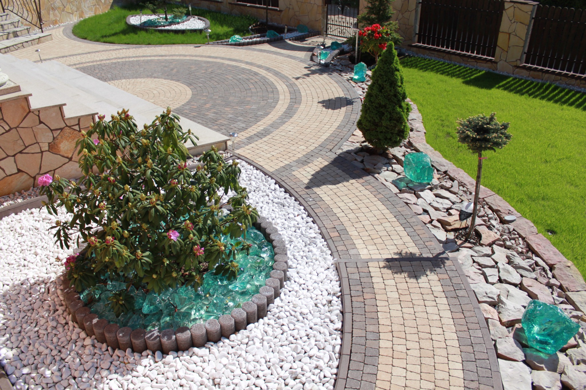 Ogród z krętą ścieżką z kostki brukowej w odcieniach beżu i szarości, ozdobiony białymi kamykami, szkłem dekoracyjnym i roślinnością, w słoneczny dzień.