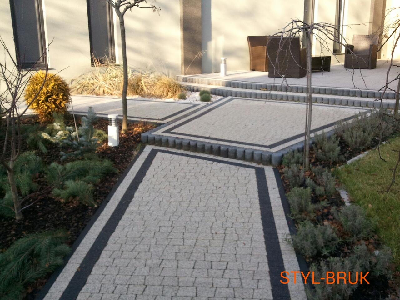 Ogród z jasnoszarą kostką z ciemnymi obramowaniami, obrzeża z szarej kostki, rośliny ozdobne wzdłuż ścieżki, nowoczesny dom w tle, oświetlenie ogrodowe.