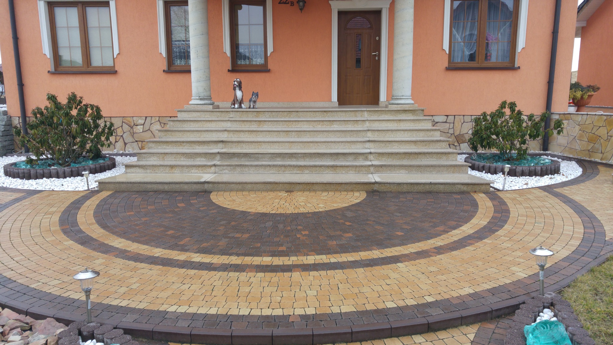 Okrągły wzór z dwukolorowej kostki brukowej przed domem z jasną elewacją, schodami i kolumnami. Na schodach dwie figurki psów, rabaty z białymi kamieniami i krzewami po bokach.