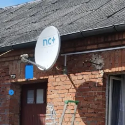 Biała antena satelitarna z logo nc+ zamontowana na ceglanej ścianie budynku, widoczna drabina oparta o ścianę, szary dach z rynnami w tle.
